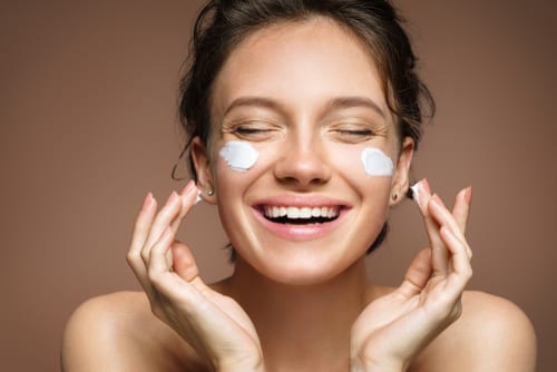 Laughing girl applying moisturizing cream on her face. Photo of young girl with flawless skin-img-blog