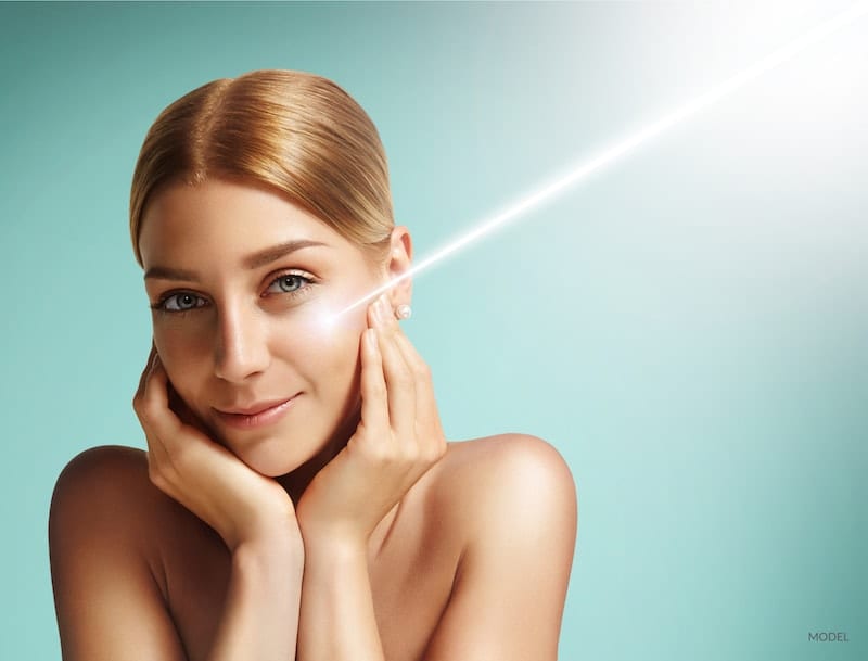 Young woman with beam of laser light shown on her cheek against teal background.