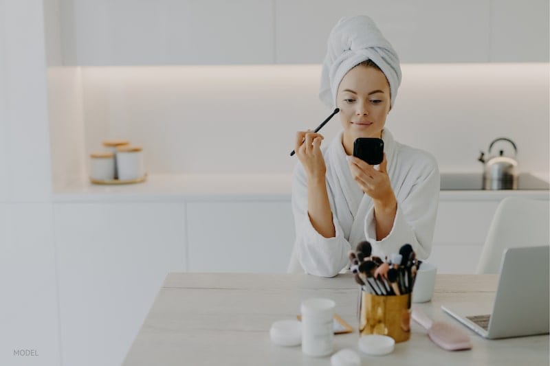 Woman applying makeup near her computer to prepare for her video conferencing call.