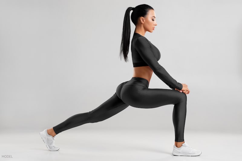 Woman doing deep lunge in black exercise clothing against a gray background.