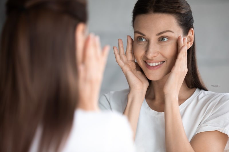 Young woman wearing white shirt looking and touching her cheeks in mirror.